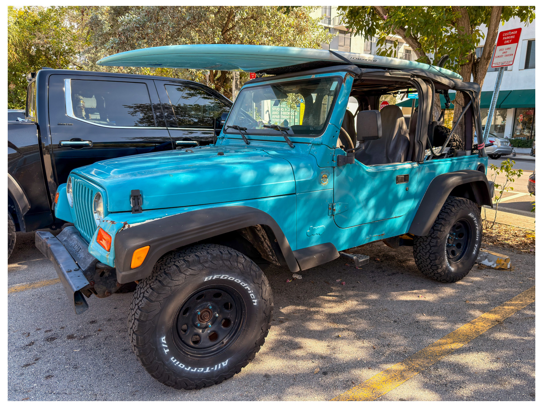 A turquoise Jeep Wrangler with the top down and a paddleboard on the roof is parked in a lot next to a black vehicle.