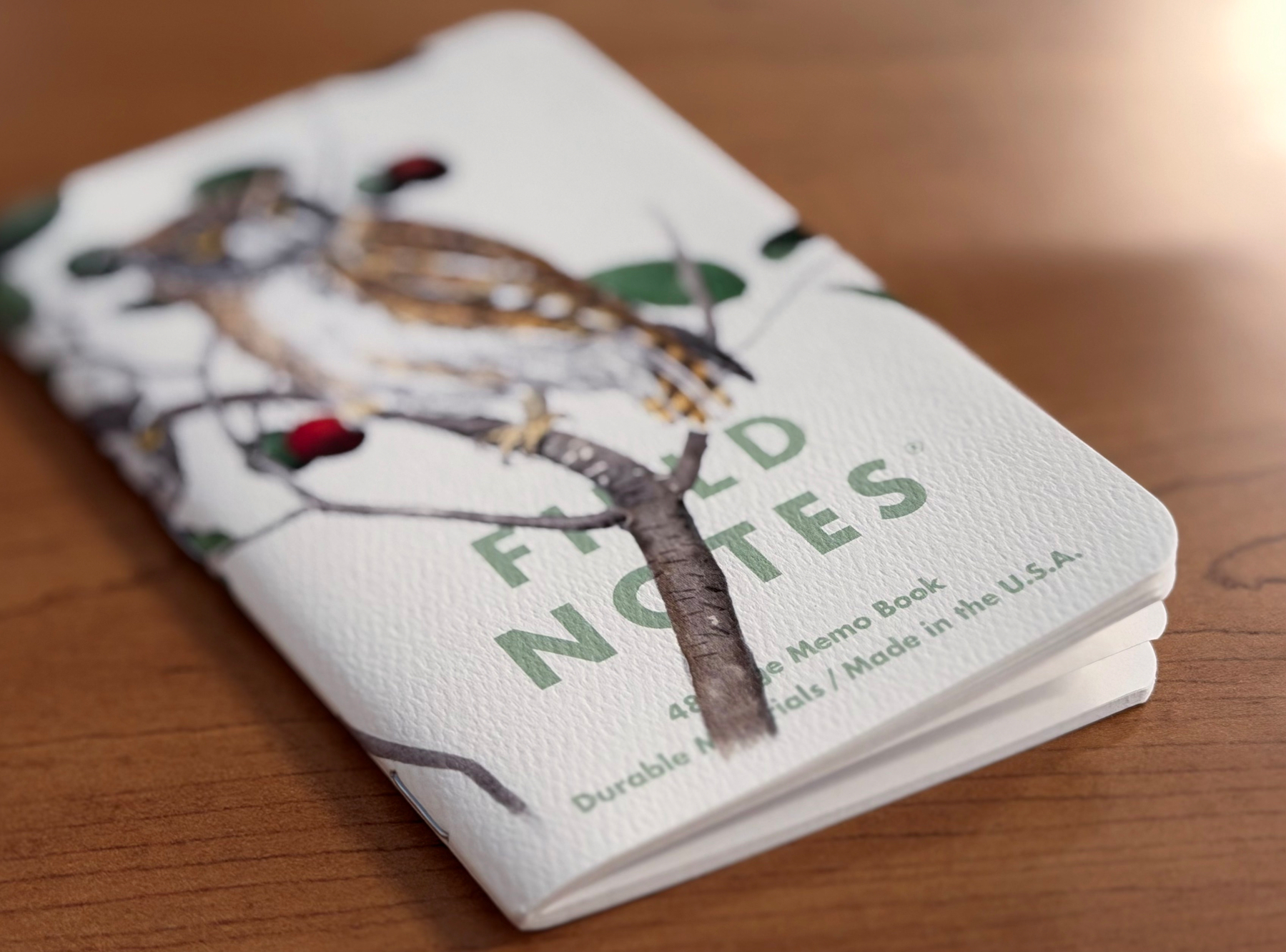A white Field Notes 48-page memo book with a painted bird perched on a branch with red berries.