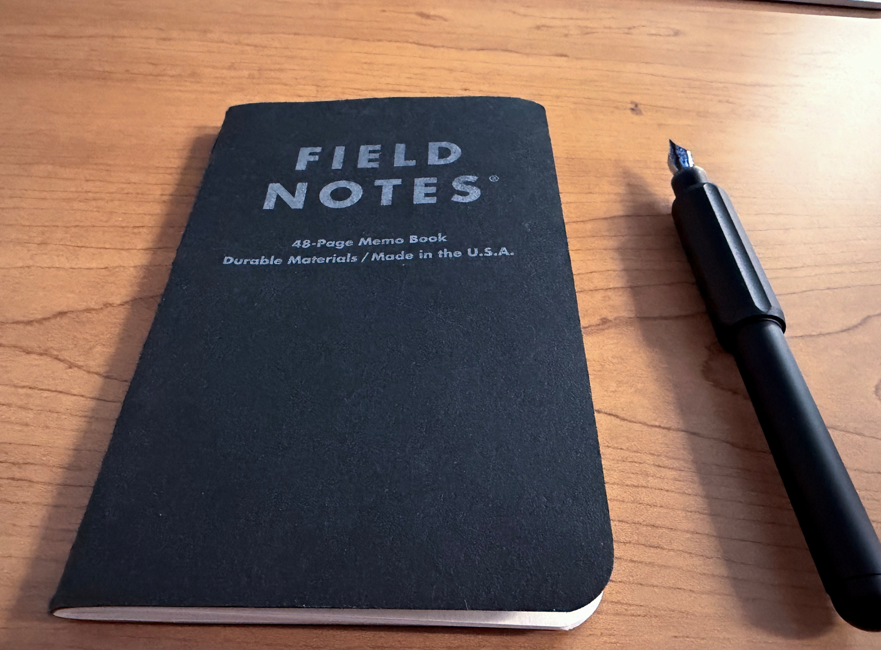 A black Field Notes 48-page memo book lies on a wooden table beside a black fountain pen.