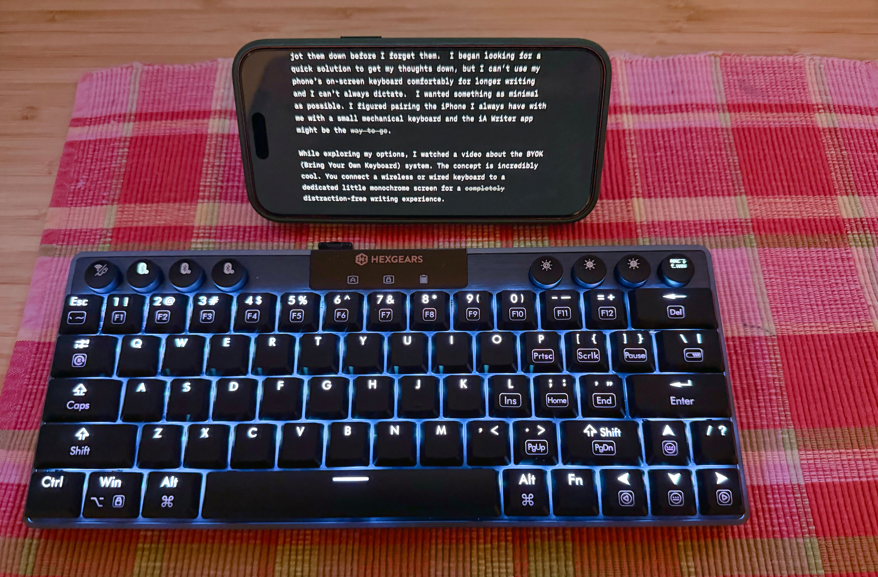 A minimal typing setup featuring a backlit Hexgears compact mechanical keyboard in front of a smartphone displaying a writing app in landscape mode, resting on a pink and red plaid cloth.
